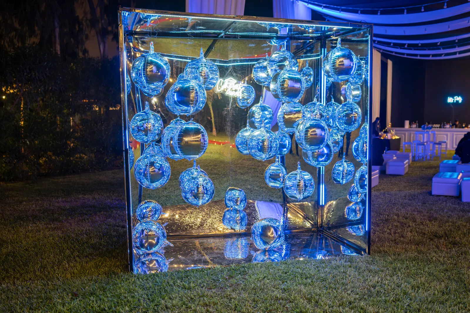 Cubo de espejos lleno de globos plateados, iluminado en azul sobre el césped
