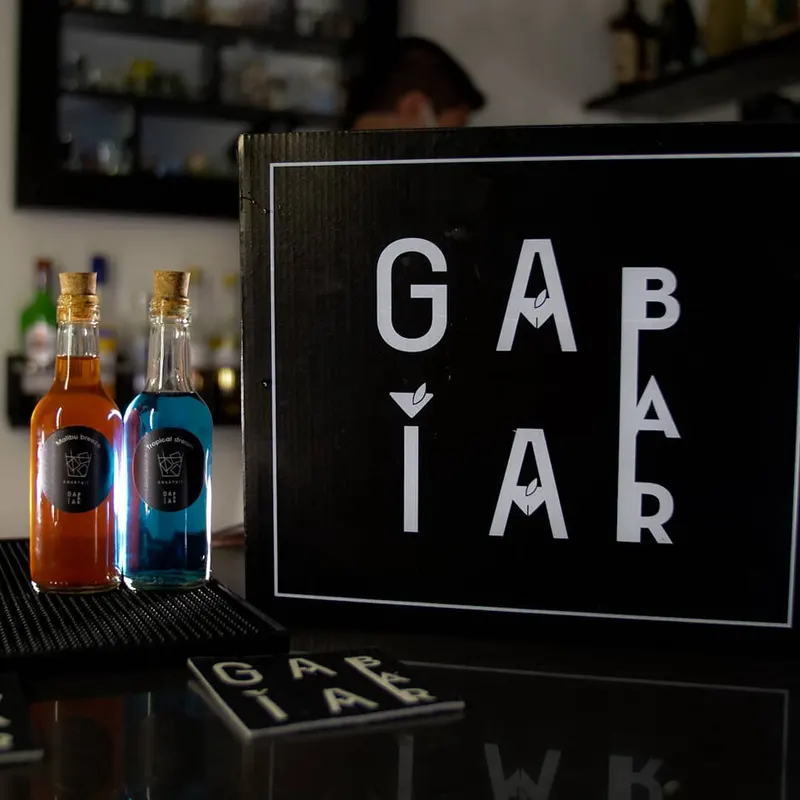 Caja negra con el logo de Gaia Bar junto a dos botellas de cóctel con etiqueta y un posavasos, sobre la barra