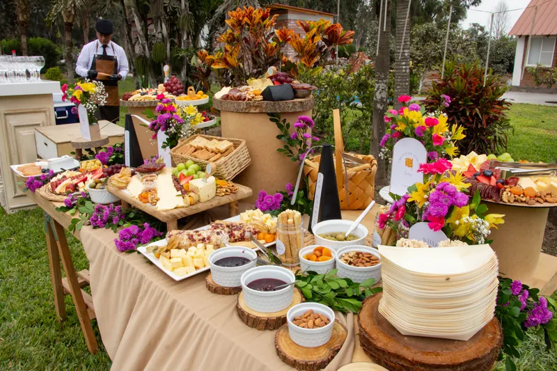 Mesa de quesos, frutas, panes y frutos secos sobre tablas de madera con flores, en un jardín