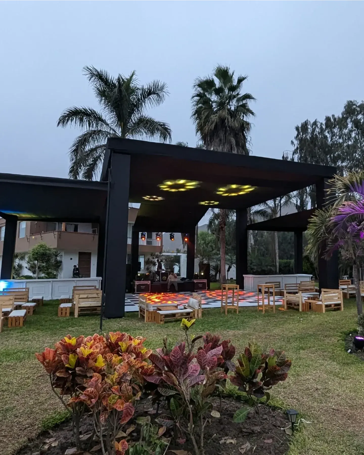 Pérgola negra con pista ajedrezada, luces de colores y mobiliario de pallets al atardecer