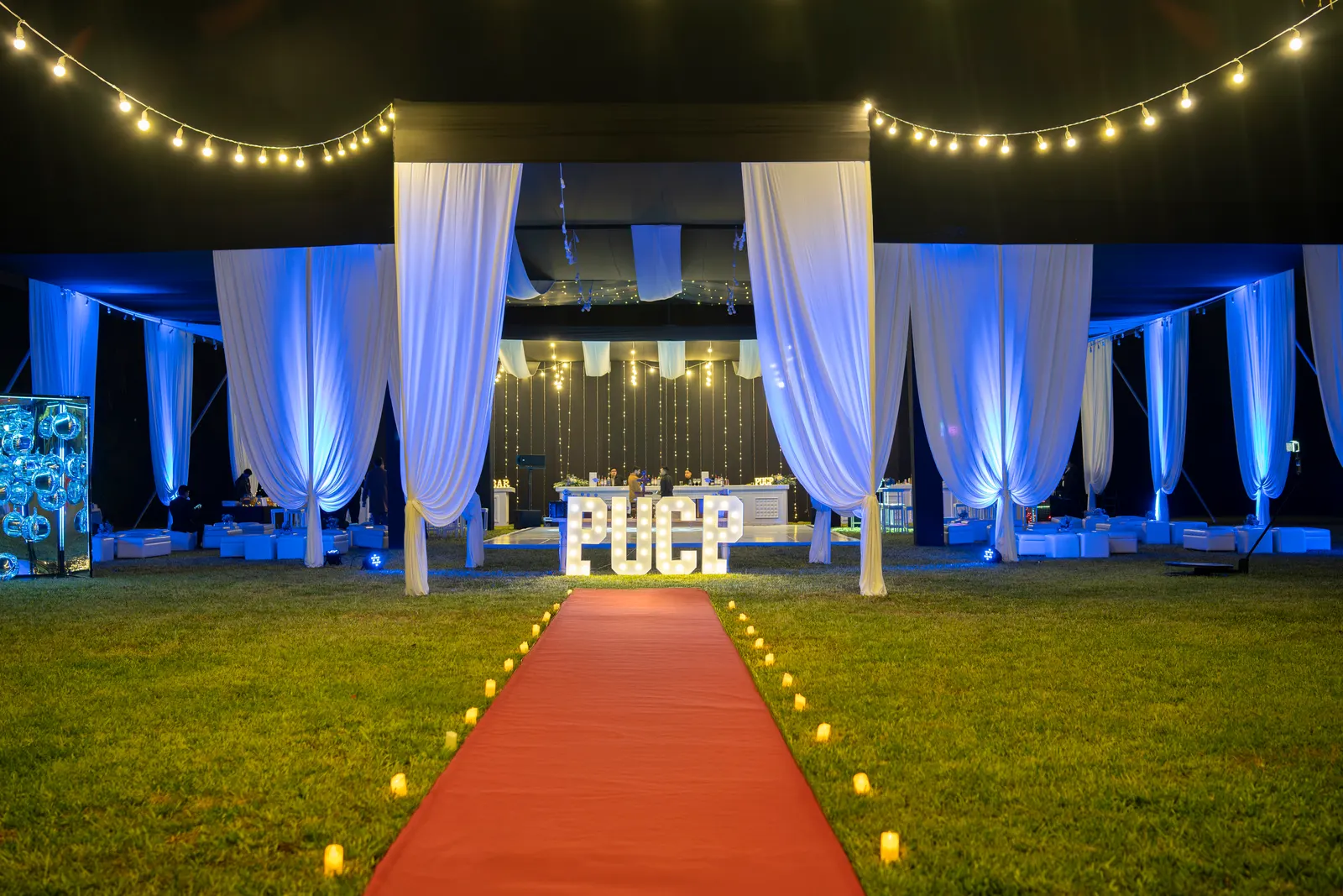 Alfombra roja con velas hacia las letras iluminadas PUCP, entre drapeados blancos y luz azul