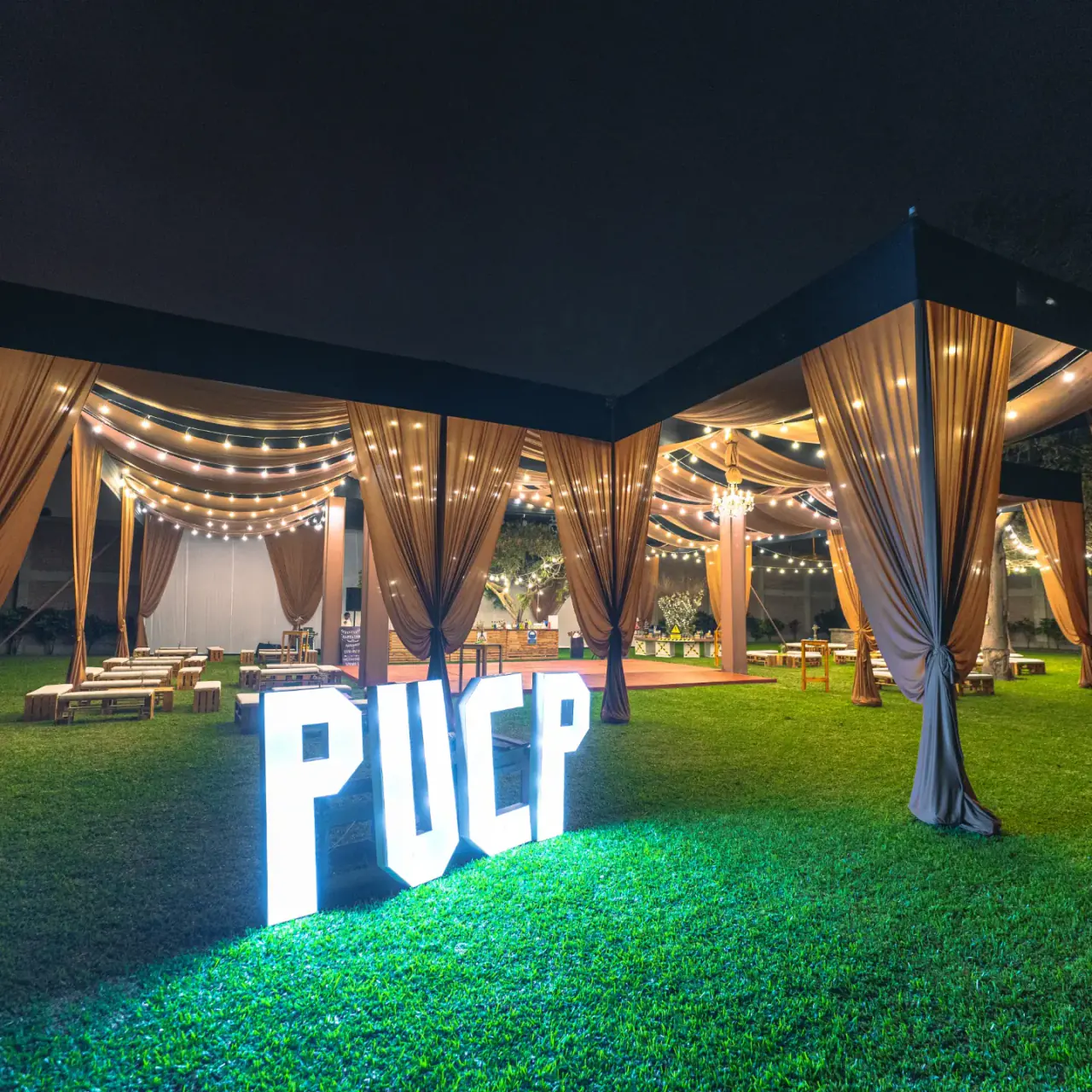 Carpa con drapeados, guirnaldas de luces y letras iluminadas PUCP sobre el césped, en una fiesta de promoción producida por Brasa