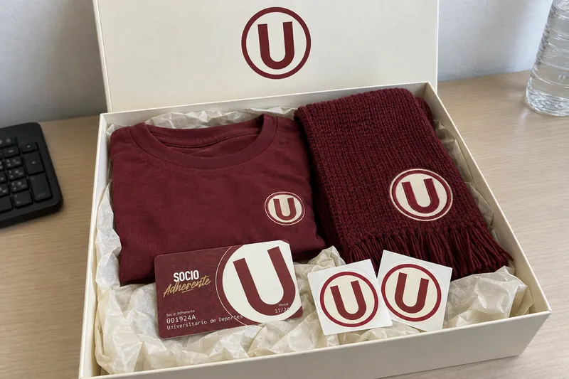 Caja crema del kit del socio de Universitario abierta: polo y bufanda granate, tarjeta de socio adherente y stickers con el escudo
