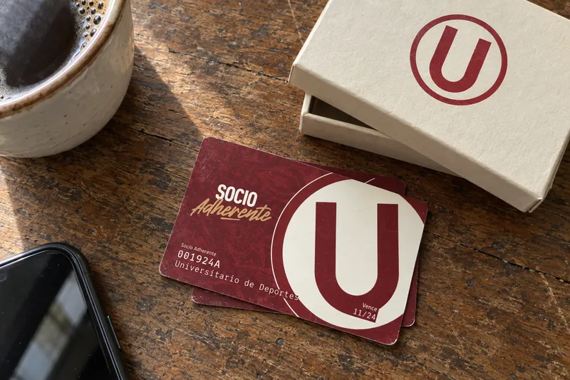 Tarjeta granate del socio adherente de Universitario con el escudo, junto a su caja crema, sobre una mesa de madera